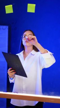 Gözlüklü, odaklanmış beyaz tenli esmer kadın panoyu tutuyor. Bayan tahtaya keçeli kalemle yazıyor. Dikey video.