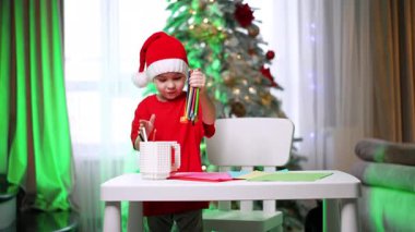 Kırmızı Noel Baba şapkalı ve tişörtlü bebek çizmeye hazırlanıyor. Sevimli çocuk büyük beyaz bir kupadan kalem topluyor. Arkaplanda Noel ağacı.