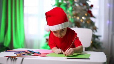 Noel Baba şapkalı sevimli beyaz çocuk kalemle resim çiziyor. Evde yaratıcı sanat hobisi yapan odaklanmış bir erkek bebek..