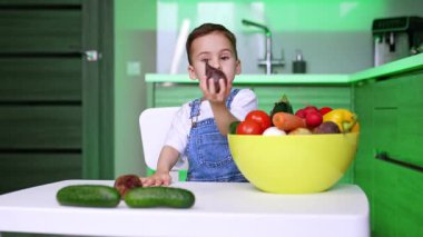 Kot pantolon giymiş sevimli bir çocuk mutfakta oturuyordu. Bebek sebze dolu bir kaseden sebze seçiyor ve masaya koyuyor..