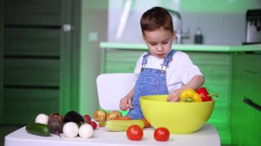 Beyaz tişörtlü ve kot pantolonlu beyaz bir bebek masada birden fazla sebzeyle duruyor. Küçük çocuk evde sebze öğreniyor..