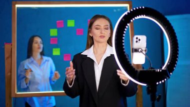 Yüzük ışığının önünde bir kadın beliriyor. Bir kadın kameraya güvenle konuşurken, diğeri arka plandaki cam bir tahtaya yapışkan notlar düzenliyor..