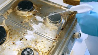 Yağlı bir mutfak ocağını temizliyorum. Mavi eldivenli eller kirli bir sobanın üzerine temizleyici sıkarak kalın yağları ve yemek artıklarını süngerle temizliyor..
