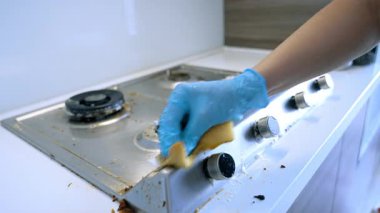 Kirli bir sobanın üstünü temizliyorum. Mavi eldiven giyen bir insan, modern bir mutfakta yağlı bir sobanın üstünü ovalayarak yemek artıklarını temizlemeye çalışır..