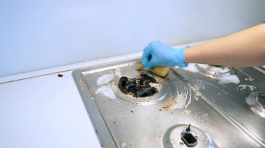 Mutfakta sobayı temizliyorum. Mavi eldiven giyen biri, kirli bir sobayı süngerle temizliyor. Parlak bir mutfakta..