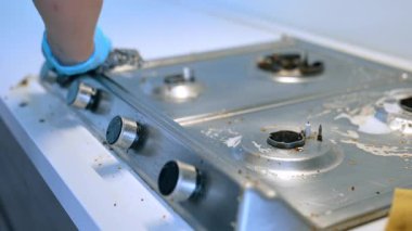 Mutfak temizleme işlemi devam ediyor. Eldiven giyen biri modern bir mutfakta kirli bir sobanın üstünü temizler. Yiyecek kalıntıları ve lekeler görülebilir..