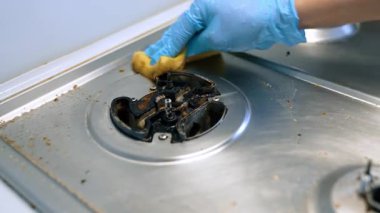 Yağlı bir sobayı fırçalıyorum. Mavi eldiven giyen bir el kirli bir sobayı süngerle ovuyor, mutfakta yağ temizleme işine odaklanıyor..