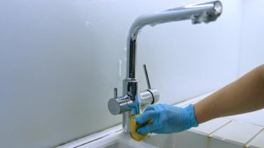 Modern bir musluğu temizliyorum. Bir insan mavi eldiven giyerken kir ve kiri temizlemek için parlak bir mutfak musluğunu ovalıyor..
