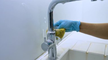 Musluğu süngerle temizliyorum. Bir insan parlak bir ortamda mavi eldiven giyerken süngerle parlak bir mutfak musluğunu ovalıyor..