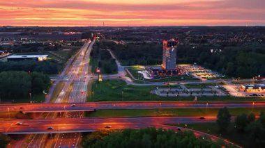 Şehir ufuk çizgisi ve otoyolun gün batımı manzarası. Canlı bir günbatımı, işlek bir otoyol ve şehir manzarasında modern bir otel üzerine renkler serpiştiriyor..