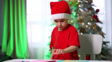 Noel Baba şapkalı çocuk tatil kartları hazırlıyor. Noel Baba şapkası takan genç bir çocuk bir masada oturup renkli tatil kartları hazırlıyor..