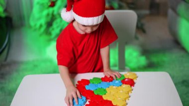 Çocuk Xmas dekorasyonu yapıyor. Noel Baba şapkası takan küçük bir çocuk masaların üzerinde renkli şekiller toplarken, yakınlarda bir festival ağacı parlıyor..