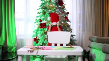 Noel ağacı dekoru ve el sanatları. Masada oturan bir çocuk süslü bir Noel ağacının yanında tatil zanaatı yapar ve siyah bir kedi seyreder..