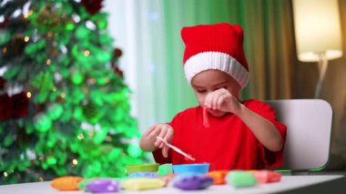 Çocuk renkli hamurla sanat yaratıyor. Kırmızı gömlekli ve şenlikli şapkalı bir çocuk tatil masasında renkli mankenlik hamuruyla oynuyor..
