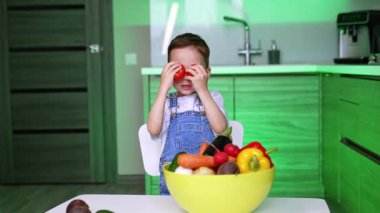 Çocuk mutfakta meyvelerle oynuyor. Genç bir çocuk gülümser, bir kase renkli meyvenin üzerine iki elma kaldırır..