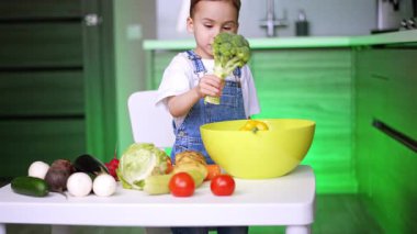 Çocuk mutfakta sağlıklı yemek hazırlıyor. Bir çocuk, sağlıklı beslenmeyi teşvik eden sebzelerle çevrili, parlak bir mutfakta brokoli tutuyor..
