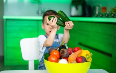 Mutfakta salatalıklı çocuk. Genç bir çocuk, parlak bir mutfakta bir kase taze sebzeyle çevrili iki salatalık sunar..