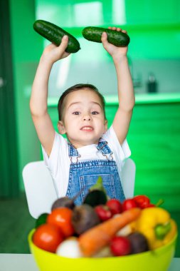 Çocuk neşeyle salatalık tutuyor. Bir çocuk renkli sebzelerle dolu parlak bir mutfakta mutlu bir şekilde salatalık yetiştiriyor..