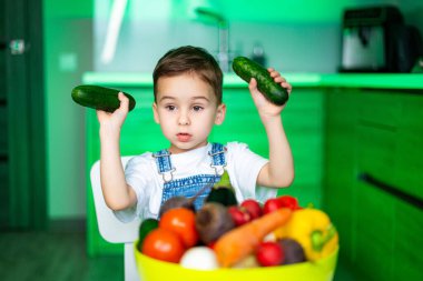Çocuk mutfakta salatalık tutuyor. Genç bir çocuk parlak bir mutfakta renkli sebzelerle dolu bir masada otururken salatalıkla oynuyor..