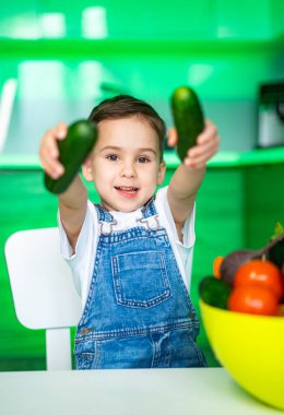 Çocuk mutlu bir şekilde salatalıkları içeride tutuyor. Genç bir çocuk, renkli meyveler ve parlak yeşil dekorlarla dolu modern bir mutfakta salatalık tutarken gülümsüyor..