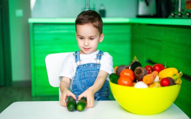 Çocuk taze sebzelere bayılıyor! Genç bir çocuk, parlak bir mutfakta renkli sebzelerle çevrili, salatalıklı beyaz bir masada oturuyor..
