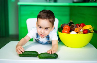 Çocuk mutfakta salatalıkla oynuyor. Bir çocuk parlak bir mutfakta renkli meyve ve sebzelerle çevrili bir masada salatalık hazırlıyor..