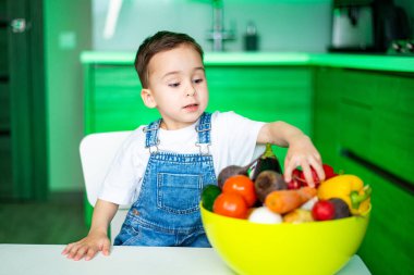Çocuk mutfakta renkli meyvelerin tadını çıkarıyor. Tulum giymiş genç bir çocuk yeşil aksanlı modern bir mutfaktaki parlak bir kaseden taze meyve seçiyor..