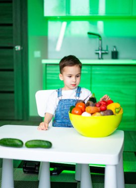 Çocuk canlı ürünleri keşfeder. Genç bir çocuk canlı bir kase meyve ve sebzeyle masada oturuyor. Merakını ve neşesini gösteriyor..