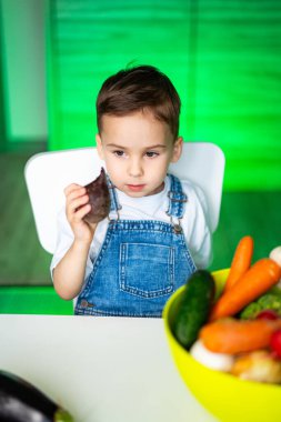 Çocuk evde sağlıklı atıştırmalıkların tadını çıkarıyor. Genç bir çocuk parlak bir mutfakta taze ürünlerle çevrili renkli bir sebze aperatifi ile masada oturuyor..