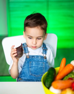 Çocuk renkli sebzelerle oynuyor. Genç bir çocuk, bir sebzeyi incelerken bir mutfakta canlı taze sebzelerle çevrili bir masada oturuyor..