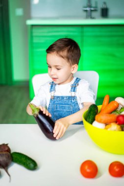 Çocuk sebzeleri öğreniyor. Genç bir çocuk taze sebzelerle bir masada oturmuş merakla patlıcanı inceliyor..