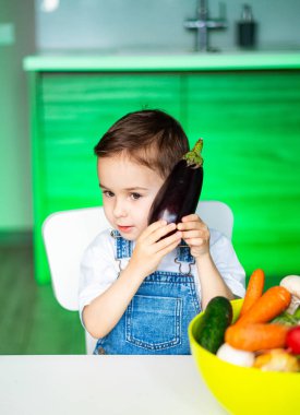Parlak mutfakta patlıcanlı bir çocuk. Genç bir çocuk, canlı bir mutfakta renkli sebzelerle dolu bir kasenin yanında şakacı bir şekilde patlıcan tutuyor..