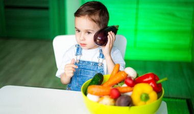Çocuk vejetaryen telefonuyla konuşuyor. Küçük bir çocuk renkli bir kase sebzeyle masada oturuyor, şakayla kulağına patlıcan dayıyor..