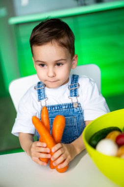 Parlak mutfakta havuçlu çocuk. Tulumlu bir çocuk, canlı meyve ve sebzelerle çevrili, parlak bir mutfakta üç havuçlu bir masada oturuyor..
