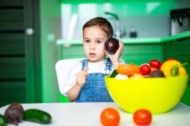 Çocuk modern bir mutfakta meyvelerden hoşlanıyor. Tulumlu genç bir çocuk renkli bir meyve kasesiyle bir masada oturuyor, parlak bir mutfakta bir tane tutuyor..