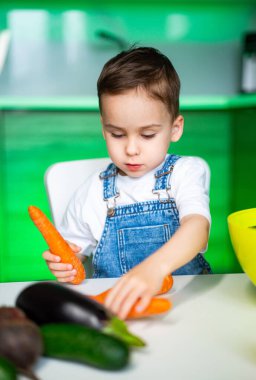 Çocuk sebze hazırlıyor. Kot pantolon giymiş genç bir çocuk, parlak bir mutfaktaki masanın üzerindeki renkli sebzeleri düzenlemeye odaklanıyor..