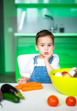 Çocuk sebzelerle oynuyor! Genç bir çocuk mutfak masasında oturmuş, modern yeşil bir dekorla çevrili renkli sebzeler ve meyveler üzerinde kafa yoruyor..