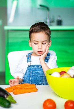 Çocuk taze ürünlerle oynuyor. Genç bir çocuk masasında oturmuş taze sebze ve meyve çeşitlerini düşünüyor. Parlak bir mutfak ortamında..