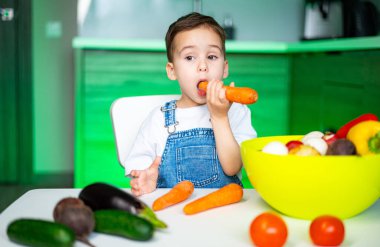 Çocuk mutfaktaki sebzelere bayılıyor. Kahverengi saçlı küçük bir çocuk, parlak bir mutfak ortamında renkli sebzeleri incelerken havucu ısırıyor..