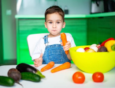 Çocuk renkli sebzelere bayılıyor. Genç bir çocuk taze sebzelerle dolu bir masada oturmuş elinde neşeli bir ifadeyle havuç tutuyor..