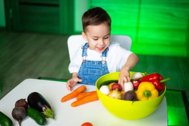 Parlak sebzelerle oynayan çocuk. Neşeli bir çocuk parlak bir odada masada otururken bir kase taze sebzeyi keşfeder..