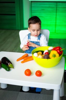 Çocuk mutlu bir şekilde sebze yiyor. Genç bir çocuk parlak bir mutfakla çevriliyken renkli meyve ve sebzeleri keşfediyor..
