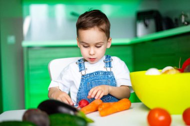 Çocuk sebzelerle oynuyor. Mavi tulumlu genç bir çocuk gün boyunca parlak bir mutfak masasında renkli sebzeleri inceliyor..