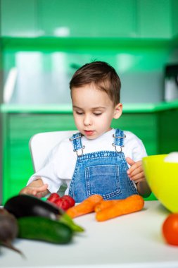 Çocuk mutfakta sebze hazırlıyor. Kot pantolonlu genç çocuk yeşil ışık ve modern dekorla dolu parlak bir mutfakta taze sebzeleri sınıflandırıyor..