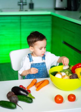 Çocuk parlak sebzelere bakıyor. Tulumlu bir çocuk, bir dizi renkli ürünle çevrili modern bir mutfakta taze sebzeleri inceliyor..