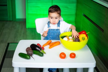 Küçük çocuk renkli sebzeleri keşfeder. Masada oturan çocuk, mutfak ortamında bir kase taze sebzeyle etkileşime girer..