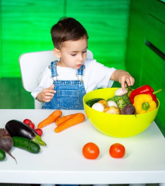 Çocuk renkli sebzeleri keşfediyor. Tulumlu bir çocuk bir kase taze sebzeyi incelerken masada daha çok sebze oluyor..