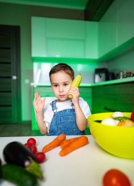 Çocuk mutfakta telefonla konuşuyor. Mutfakta küçük bir çocuk kulağına salatalık tutar, taze sebzeler ve meyvelerle çevrili..