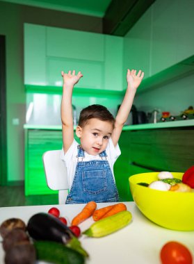 Çocuk sebze pişirmeyi seviyor. Genç bir çocuk, taze sebzeler ve yemek malzemeleriyle çevrili mutfak masasında heyecanlı bir şekilde ellerini kaldırır..