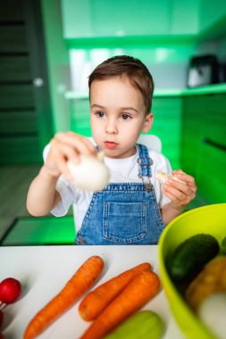 Çocuk mutfakta taze yemek pişiriyor. Kot pantolonlu bir çocuk, mutfak masasında otururken, taze sebzelerle çevrili bir yumurtayı inceliyor..